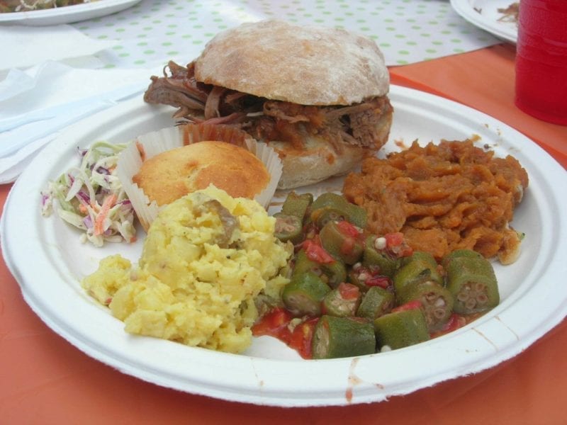 Plate of pulled pork on a bun, potato salad, corn bread muffin, coleslaw, and mashed sweet potatoes.