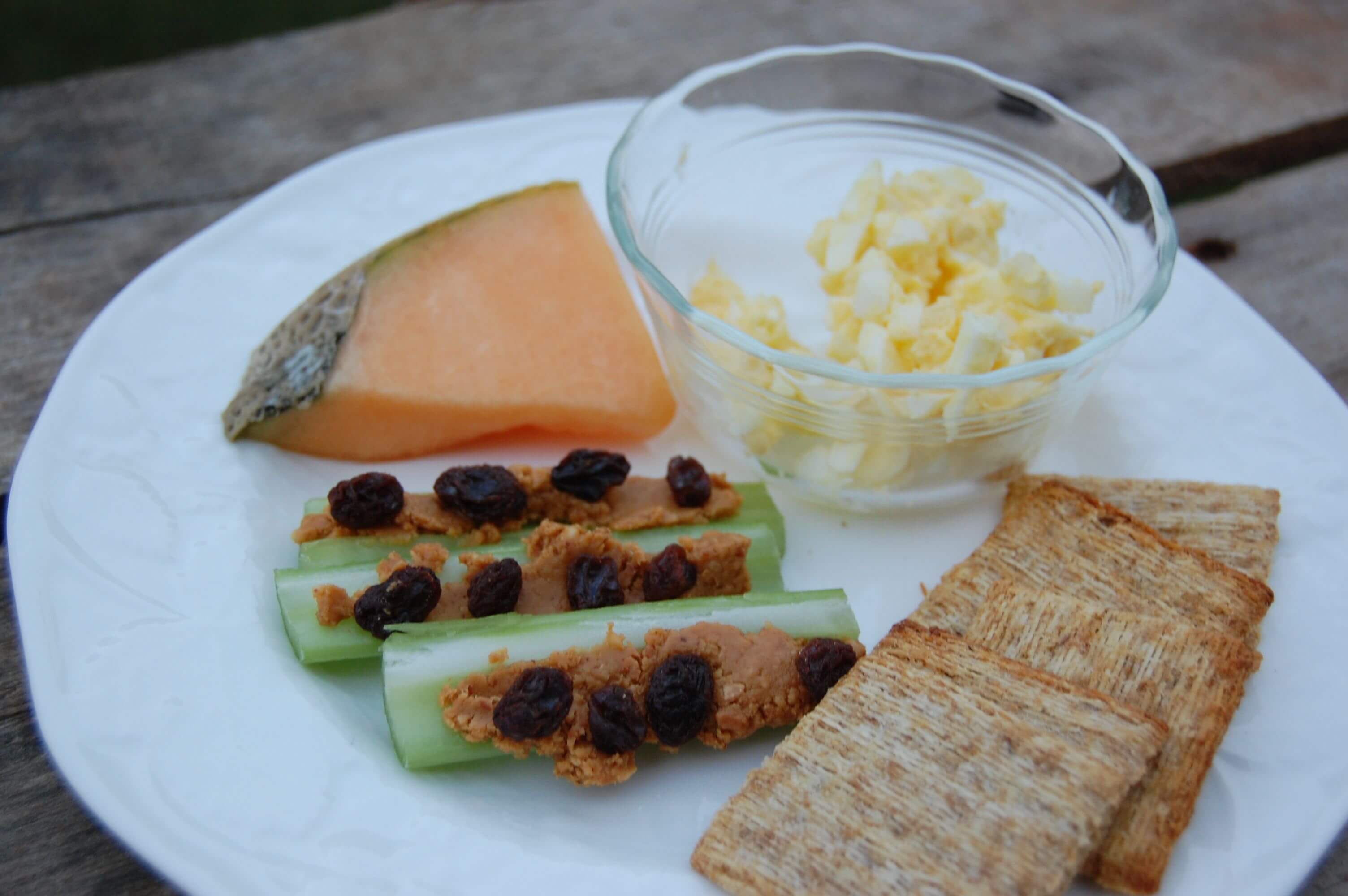 A plate of egg salad, crackers, celery with peanut butter & raisins, and a slice of cantaloupe.